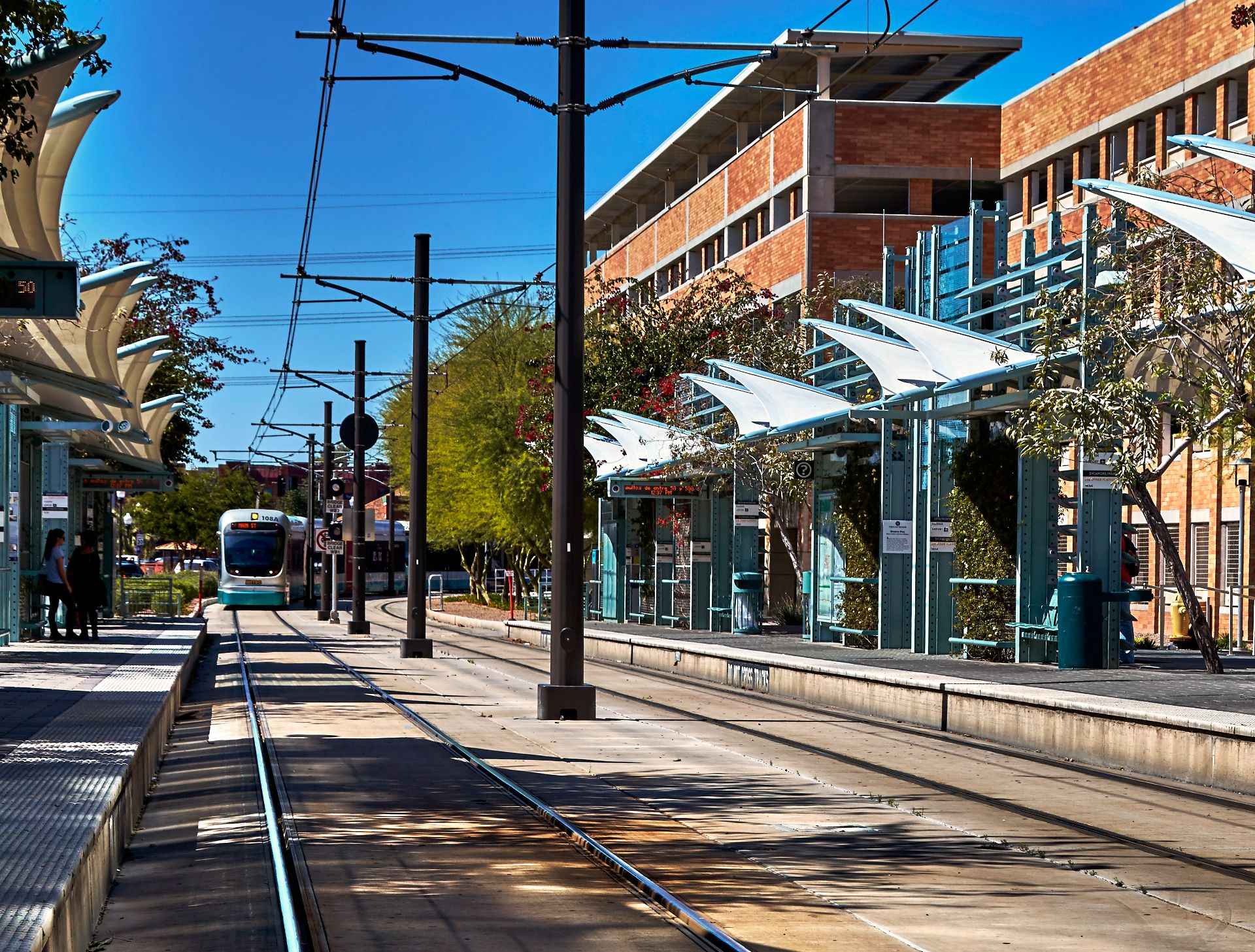A light rail train approaching a modern station with glass canopies on a sunny day.