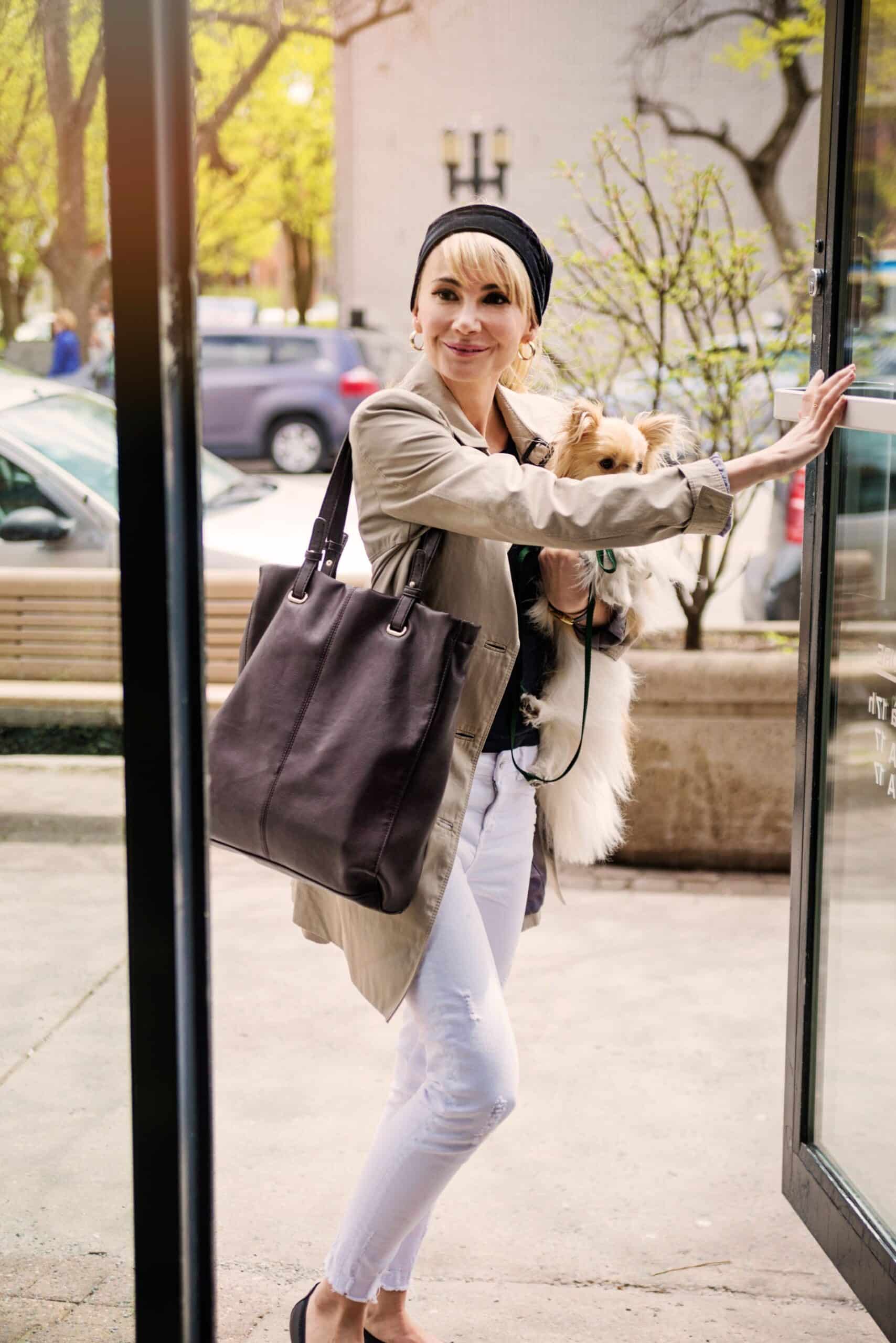 A person stepping out of a building holding a small dog and a bag.