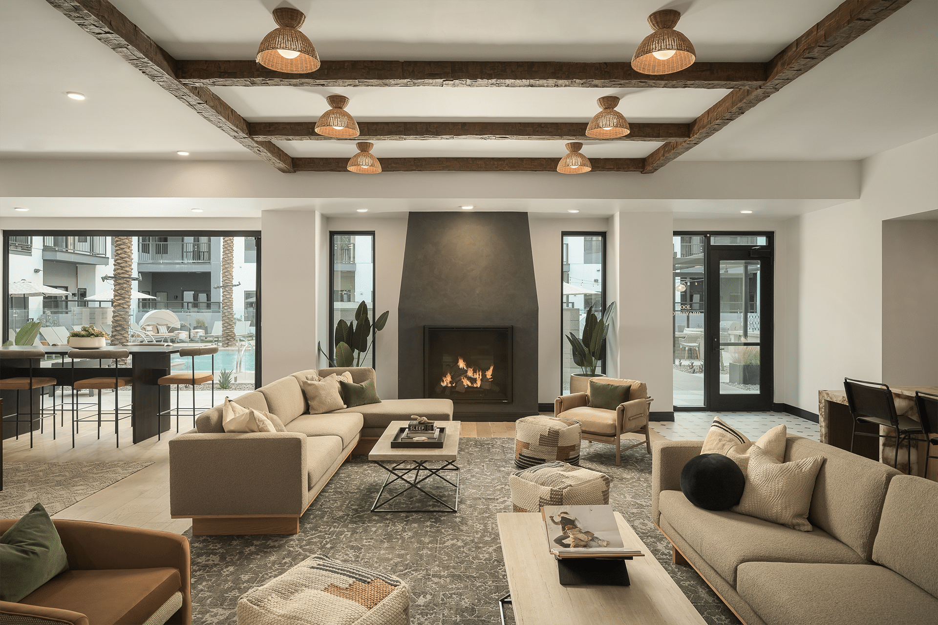 Spacious resident lounge with neutral tones, a central fireplace, exposed wooden beams, and large windows.