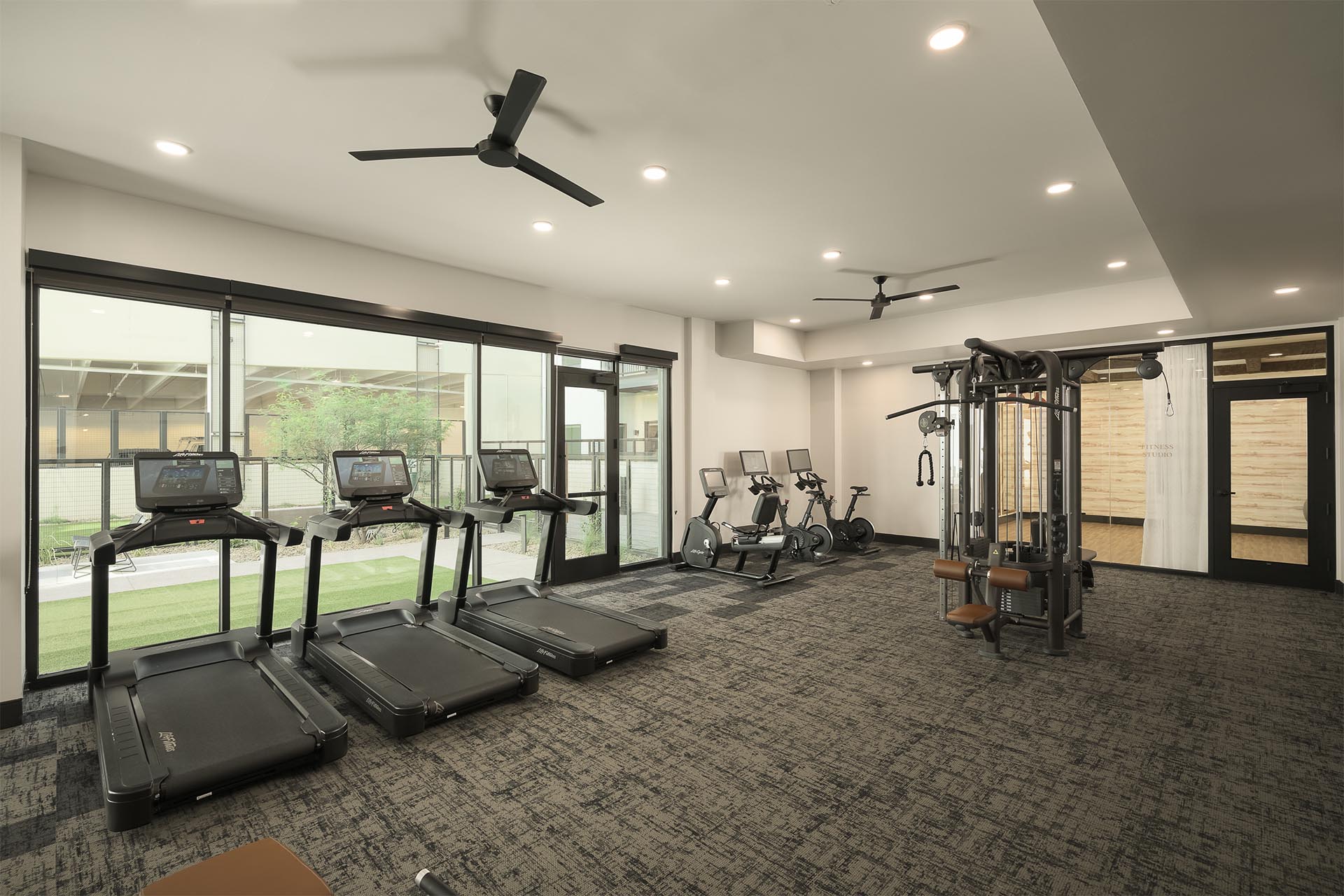 Fitness center with treadmills, elliptical machines, and a multi-station weight machine by large windows.
